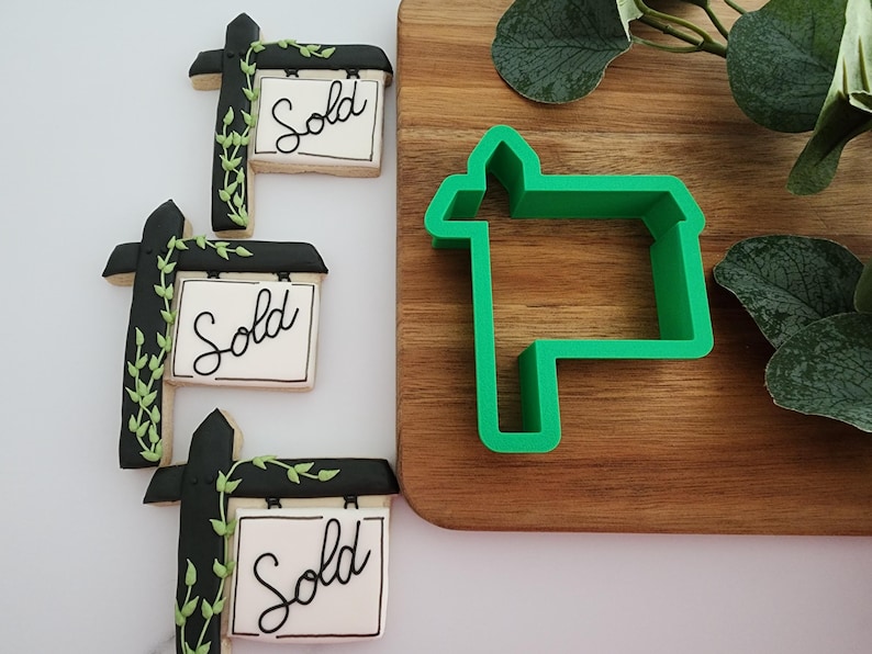 Puede incluir: Tres galletas decoradas con glaseado negro y verde, con forma de casa con un cartel que dice "Sold". Un cortador de galletas de pl&aacute;stico verde con forma de casa con un cartel est&aacute; sobre una tabla de cortar de madera.