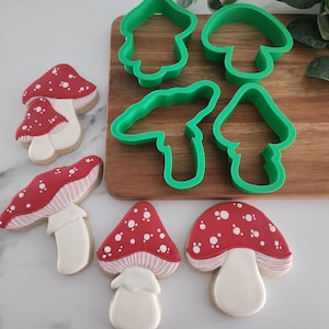 May include: An assortment of mushroom-shaped cookies with red caps and white polka dots, alongside green plastic cookie cutters in various mushroom shapes. The cookies have white stems and are arranged on a wooden board.