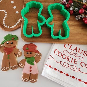 May include: Two gingerbread cookies decorated with icing and red and green accents. The cookies are shaped like people wearing cowboy hats. The cookies are next to two green plastic cookie cutters shaped like people. A white tea towel with red text that reads "Claus Cookie Co." is in the background.