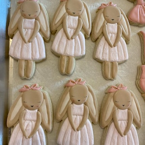 May include: Decorated sugar cookies in the shape of bunnies wearing dresses. The cookies are light brown with pink bows and dresses. The bunny faces have closed eyes and small black details. The cookies are arranged on a baking sheet.