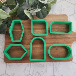 May include: Six green plastic cookie cutters on a wooden surface. The cutters are shaped like a rectangle, oval, hexagon, arch, and two different variations of a rectangle.