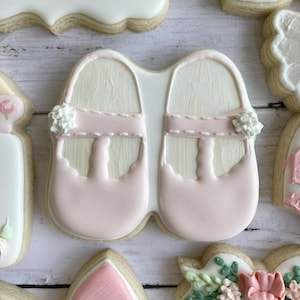 May include: Two pink and white sugar cookies decorated to look like baby shoes. The cookies have a white woodgrain design and pink straps with white flowers.