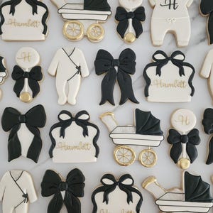 May include: A set of 12 black and white sugar cookies decorated with gold accents. The cookies feature various baby-themed designs, including a baby carriage, a rattle, a bow, and a onesie. The cookies are personalized with the name "Hamlett".