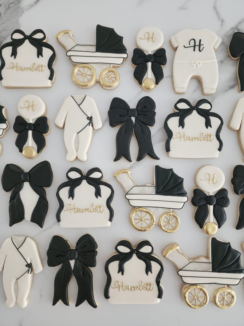 May include: A set of 12 black and white sugar cookies decorated with gold accents. The cookies feature various baby-themed designs, including a baby carriage, a rattle, a bow, and a onesie. The cookies are personalized with the name "Hamlett".