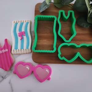 May include: Cookies and cookie cutters in a summer theme. The cookies include a pink swimsuit with "Barbie" text, a beach towel, and heart-shaped sunglasses. Green plastic cookie cutters are also present.