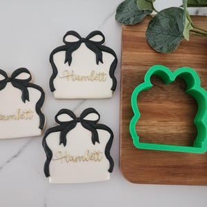May include: Three white sugar cookies decorated with black ribbon bows and the name "Hamlett" in gold lettering. A green plastic cookie cutter in the shape of a present box is on a wooden surface.