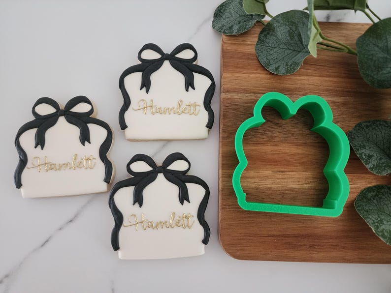May include: Three white sugar cookies decorated with black ribbon bows and the name "Hamlett" in gold lettering. A green plastic cookie cutter in the shape of a present box is on a wooden surface.