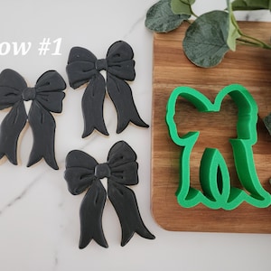 May include: Three black bow-shaped cookie cutters and a green plastic bow-shaped cookie cutter on a wooden surface. The text "bow #1" is written in white on a white background.