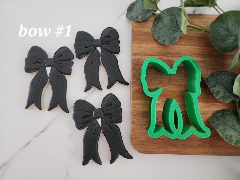 May include: Three black bow-shaped cookie cutters and a green plastic bow-shaped cookie cutter on a wooden surface. The text "bow #1" is written in white on a white background.