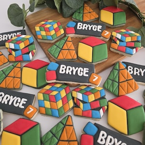 May include: A collection of decorated sugar cookies shaped like colorful puzzle cubes, pyramids, and rectangular plaques with the name Bryce and the number 7 written in white icing. The cookies feature vibrant red, yellow, blue, green, and orange colors.