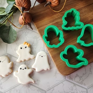May include: Five white ghost-shaped cookies with black eyes and smiles, and four green plastic ghost-shaped cookie cutters on a wooden surface.