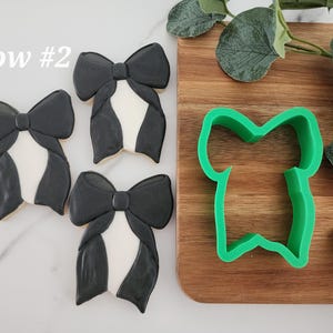 May include: Three black and white bow-shaped cookies with a green plastic cookie cutter in the shape of a bow. The text "bow #2" is visible in the top left corner of the image.