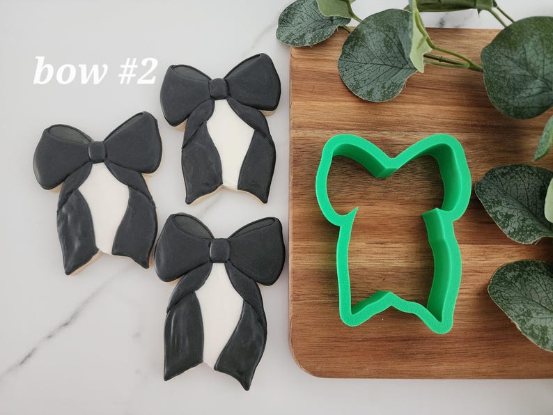 May include: Three black and white bow-shaped cookies with a green plastic cookie cutter in the shape of a bow. The text "bow #2" is visible in the top left corner of the image.
