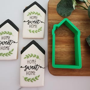 Puede incluir: Tres galletas blancas decoradas con hojas verdes y letras negras que dicen "Home sweet home". Un cortador de galletas de pl&aacute;stico verde en forma de casa est&aacute; sobre una tabla de cortar de madera.