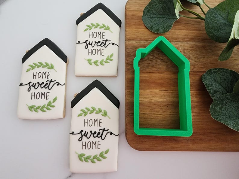 Puede incluir: Tres galletas blancas decoradas con hojas verdes y letras negras que dicen "Home sweet home". Un cortador de galletas de pl&aacute;stico verde en forma de casa est&aacute; sobre una tabla de cortar de madera.