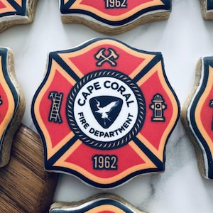 May include: Close-up of several sugar cookies decorated with the Cape Coral Fire Department emblem. The cookies feature a red and black design with the text "CAPE CORAL FIRE DEPARTMENT" and the year "1962". The emblem includes a bird, ladder, axe, and fire hydrant.