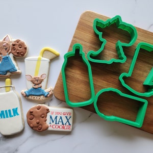 May include: A set of cookie cutters featuring a milk bottle, a glass of milk, a cookie, and a mouse. The cutters are green and are on a wooden cutting board. The text "If you give Max a cookie" is on a cookie.