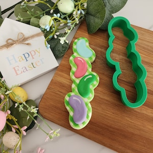 May include: A green plastic cookie cutter shaped like a row of jelly beans. A decorated cookie with four jelly bean shapes in different colors is next to the cutter. The cookie is on a wooden cutting board. The background includes a sign that says "Happy Easter" and a sprig of greenery with small eggs.