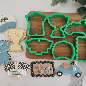 May include: An assortment of racing-themed cookies and green cookie cutters. The cookies include a trophy, race car, checkered flags, and a racing uniform. The cookie cutters are in the shape of a shirt, trophy, race car, and more. The sign says "Fast & Curious".