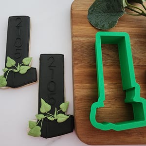 Puede incluir: Dos cortadores de galletas negros en forma de un edificio alto con una planta que crece desde la parte superior. Los cortadores de galletas est&aacute;n sobre una superficie blanca. Un cortador de galletas de pl&aacute;stico verde de la misma forma est&aacute; sobre una tabla de cortar de madera.