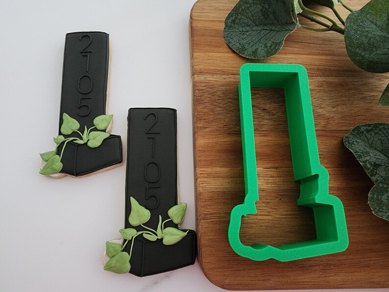 Puede incluir: Dos cortadores de galletas negros en forma de un edificio alto con una planta que crece desde la parte superior. Los cortadores de galletas est&aacute;n sobre una superficie blanca. Un cortador de galletas de pl&aacute;stico verde de la misma forma est&aacute; sobre una tabla de cortar de madera.