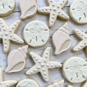 May include: A collection of 10 decorated sugar cookies in the shape of seashells, sand dollars, and starfish. The cookies are white with pink accents and have a textured surface.