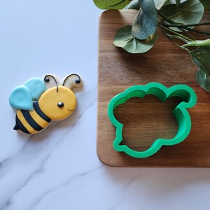 May include: A green plastic cookie cutter shaped like a bee. The cutter is on a wooden surface. A yellow and black bee cookie is on a white surface.