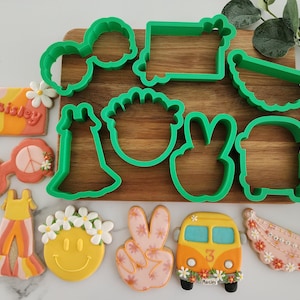 May include: A set of green plastic cookie cutters in various shapes, including a peace sign, a flower, a dress, a van, and a headband. The cutters are arranged on a wooden surface with several decorated cookies, including a peace sign cookie, a smiley face cookie, a flower cookie, and a van cookie.