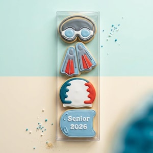 May include: A clear plastic box contains four decorated sugar cookies. The cookies are shaped like swimming goggles, flippers, a baseball, and a banner that reads "Senior 2026". The cookies have blue, red, and white icing.