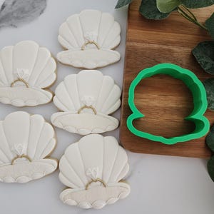 May include: Six white seashell-shaped cookies with diamond ring decorations. A green seashell-shaped cookie cutter sits on a wooden cutting board. Green leaves are in the background. The cookies are likely for a wedding or engagement.