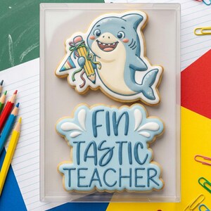 May include: Two decorated sugar cookies in a clear box. One cookie is a smiling cartoon shark holding a pencil and wearing a party hat. The other cookie says "Fin Tastic Teacher" in blue icing.