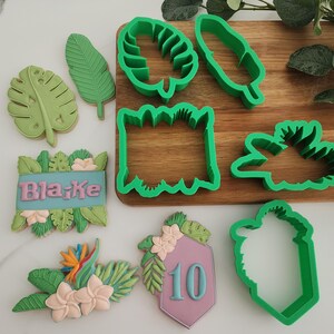 May include: Green plastic cookie cutters in various tropical leaf and floral shapes, including a banner that reads "Blake" and a sign with the number "10".