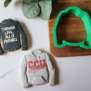 May include: Two decorated sugar cookies. One cookie is shaped like a leather jacket with the text "Through love, all is possible". The other cookie is shaped like a hoodie with the text "Champions CCU Surfball".