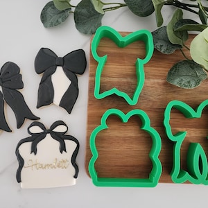 May include: Four green plastic cookie cutters in the shape of bows and a gift box. The cutters are on a wooden surface with a green leafy plant in the background.