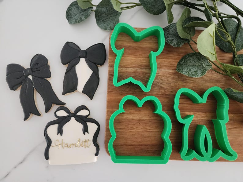 May include: Four green plastic cookie cutters in the shape of bows and a gift box. The cutters are on a wooden surface with a green leafy plant in the background.