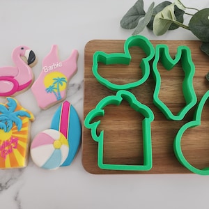 May include: Cookies and cookie cutters with a summer theme. The cookies include a pink flamingo, a swimsuit with "Barbie" text, a palm tree with "Birthday Girl" text, a beach ball, and a surfboard. Green plastic cookie cutters are also present.