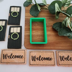 Puede incluir: Tres galletas decoradas con forma de puertas con coronas, un cortador de galletas verde y tres galletas con forma de felpudos con la palabra "Bienvenido" escrita en ellas.