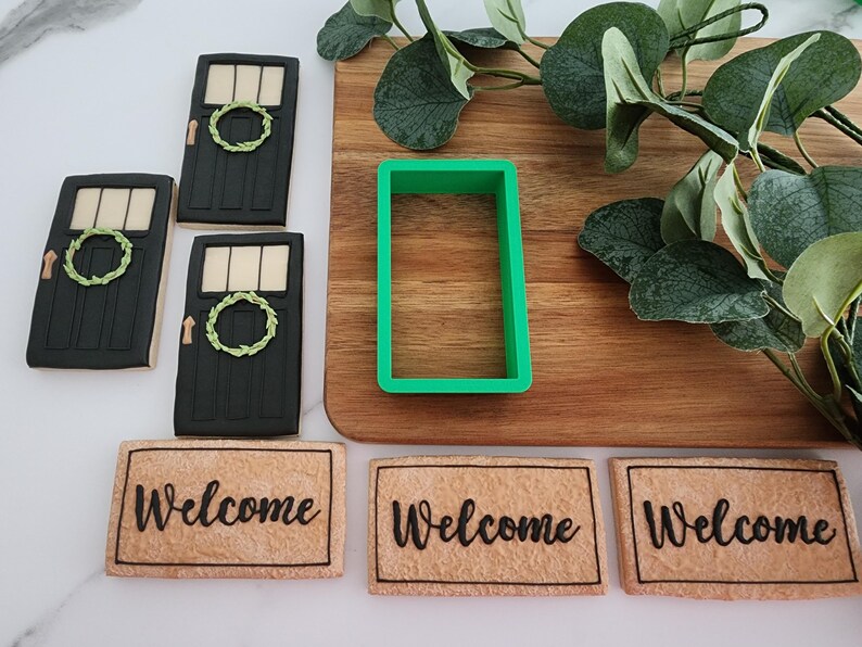 Puede incluir: Tres galletas decoradas con forma de puertas con coronas, un cortador de galletas verde y tres galletas con forma de felpudos con la palabra "Bienvenido" escrita en ellas.