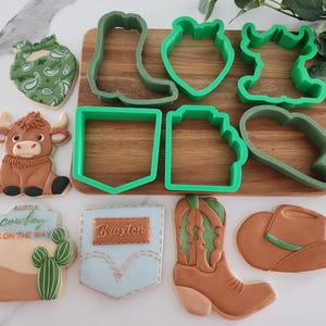 May include: A collection of cowboy-themed cookies and cookie cutters. The cookies include a bull, bandana, cowboy boot, and hat. Green plastic cookie cutters are in the shapes of a boot, heart, and other western designs. A wooden cutting board is in the background.