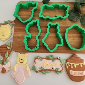 May include: Green plastic cookie cutters shaped like a beehive, a Winnie the Pooh character, a honey pot, and a sign with leaves.  Decorated cookies include a honey pot, a Winnie the Pooh character, a beehive, a honey dipper, and a sign that says "Alyana".