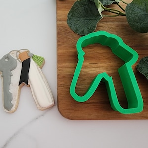 Puede incluir: Un cortador de galletas de pl&aacute;stico verde en forma de casa con un agujero de cerradura. El cortador de galletas est&aacute; sobre una superficie de madera junto a una galleta decorada con forma de llave.