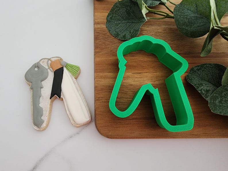 Puede incluir: Un cortador de galletas de pl&aacute;stico verde en forma de casa con un agujero de cerradura. El cortador de galletas est&aacute; sobre una superficie de madera junto a una galleta decorada con forma de llave.