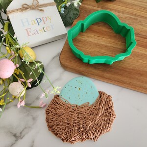 May include: A blue and gold speckled chocolate egg in a brown chocolate nest. The egg is surrounded by a green plastic cookie cutter in the shape of a bird.