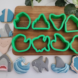 May include: A collection of shark-themed cookies and green cookie cutters on a wooden cutting board. The cookies feature shark designs, including a shark fin, a shark's mouth, and a wave. The green cookie cutters are in various shark-related shapes.