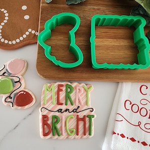 May include: Green plastic cookie cutters shaped like a Christmas tree and a rectangle with a scalloped edge.  A pink, green, and red cookie decorated with icing and the words "Merry and Bright" is on a white surface.