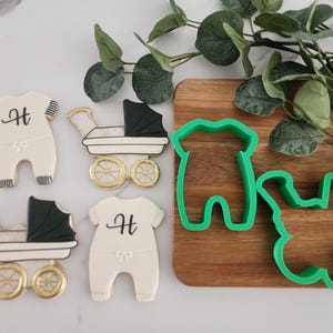 May include: Four cookie cutters: two baby onesies, one stroller, and one baby foot. The onesies have the letter "H" on them. The cookie cutters are green and on a wooden surface.
