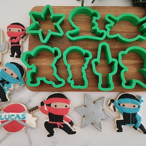May include: A collection of ninja-themed cookies and green cookie cutters on a wooden board. The cookies feature ninja characters in red and blue, along with a ninja star and a sword. One cookie has the name "LUCAS" on it.