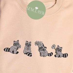 Op de afbeelding: Een beige sweatshirt met een geborduurd ontwerp van vijf wasberen die bloemen vasthouden. De wasberen zijn grijs met zwarte ogen en neuzen. De bloemen zijn wit met gele centra. De tekst "SENCLASSY" is in het groen geborduurd op de linkerbovenhoek van het sweatshirt.
