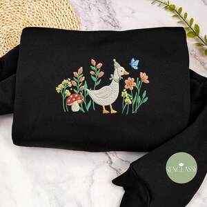 May include: Black sweatshirt with a white goose embroidered on the front. The goose is wearing a small hat and is surrounded by flowers and a mushroom.