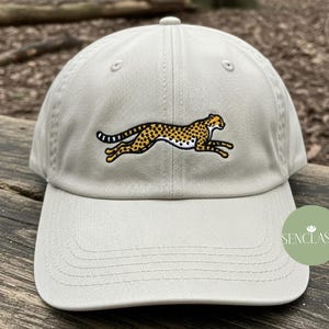 May include: A light grey baseball cap with an embroidered cheetah design in yellow, black, and white. The cheetah is shown running. The cap has a curved peak and a classic six-panel design.